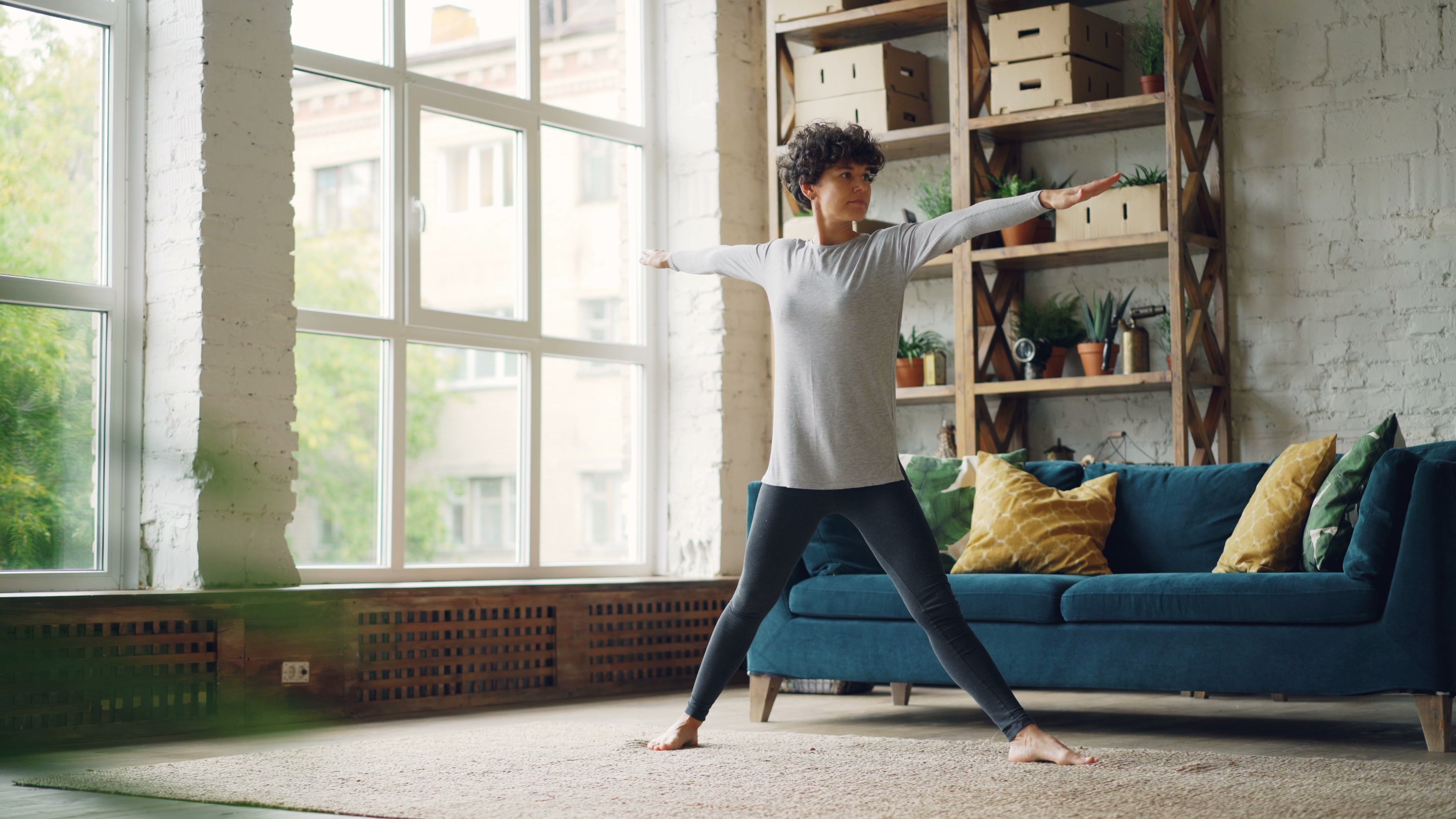 Person practising a gentle standing pose in a bright living room with a teal sofa, showing how you can build a fitness habit at home without equipment