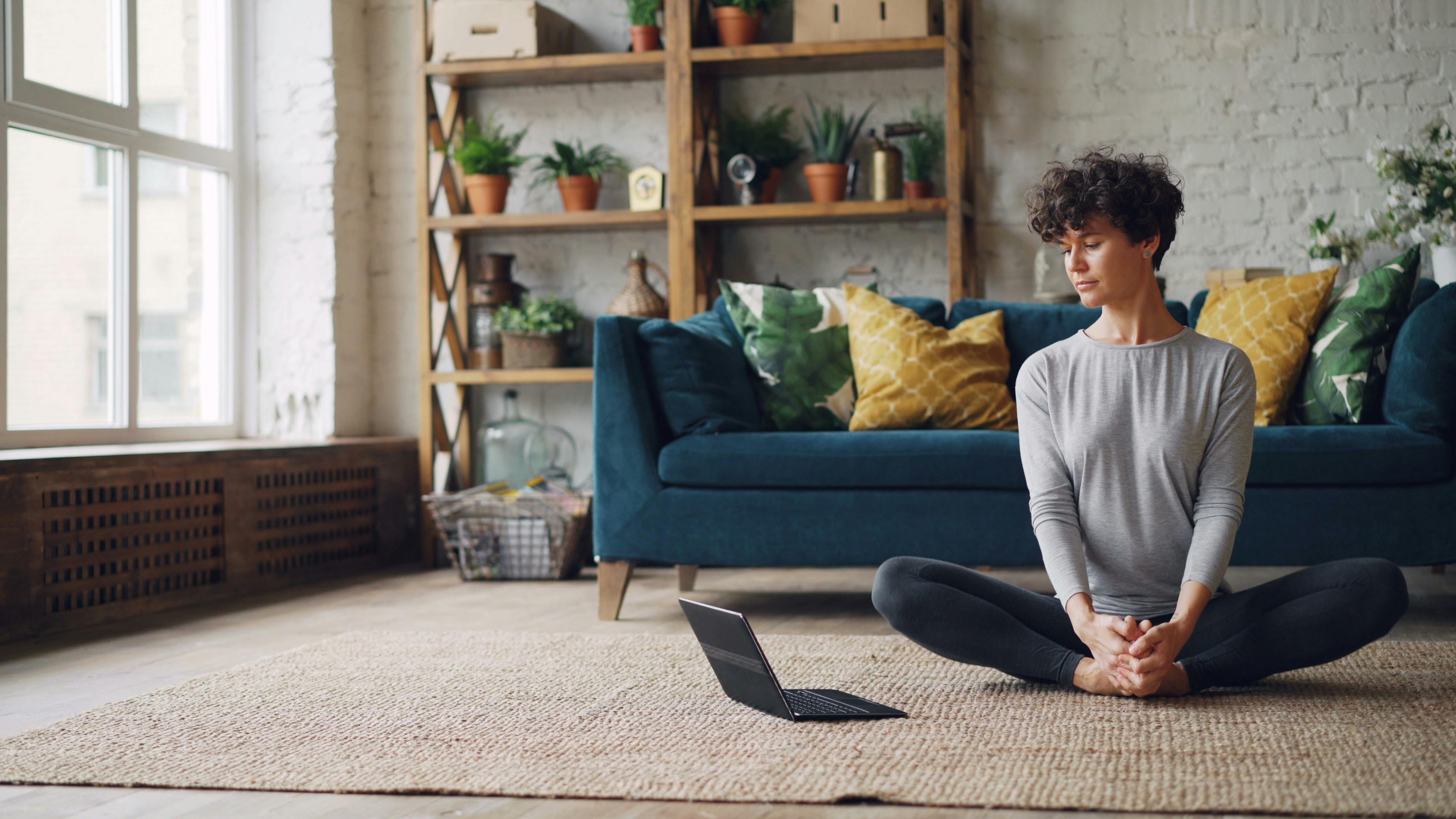 A woman meditates in a bright, cozy living room with plants and a sofa.