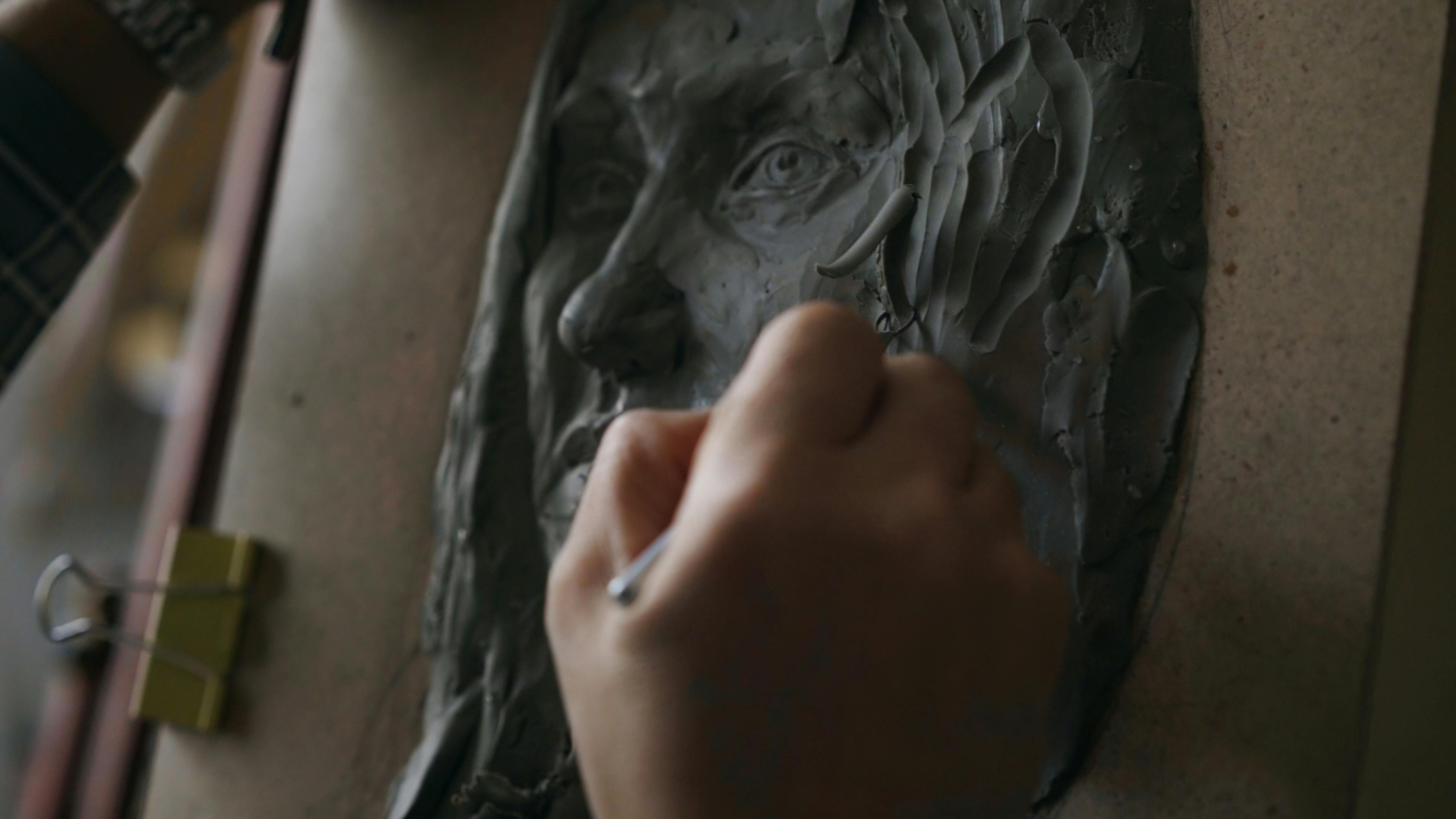 Close-up of a hand sculpting a clay face on an artist's easel indoors.