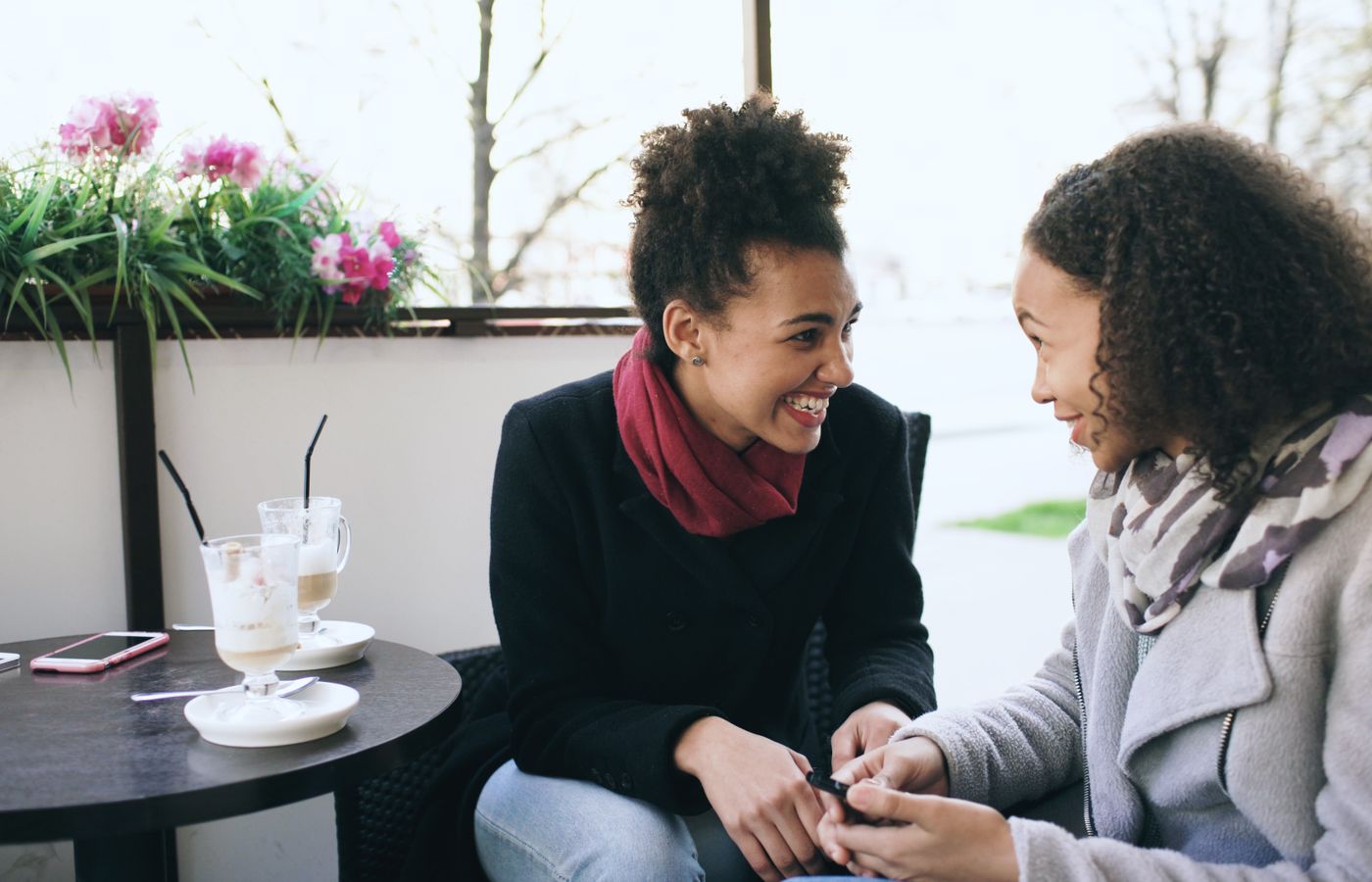 Two women smiling and chatting over a coffee at an outdoor cafe table.