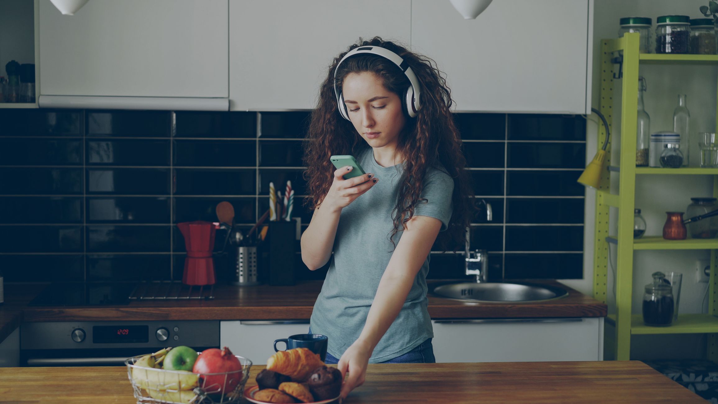 A woman in a kitchen listening to music with headphones while using her smartphone.