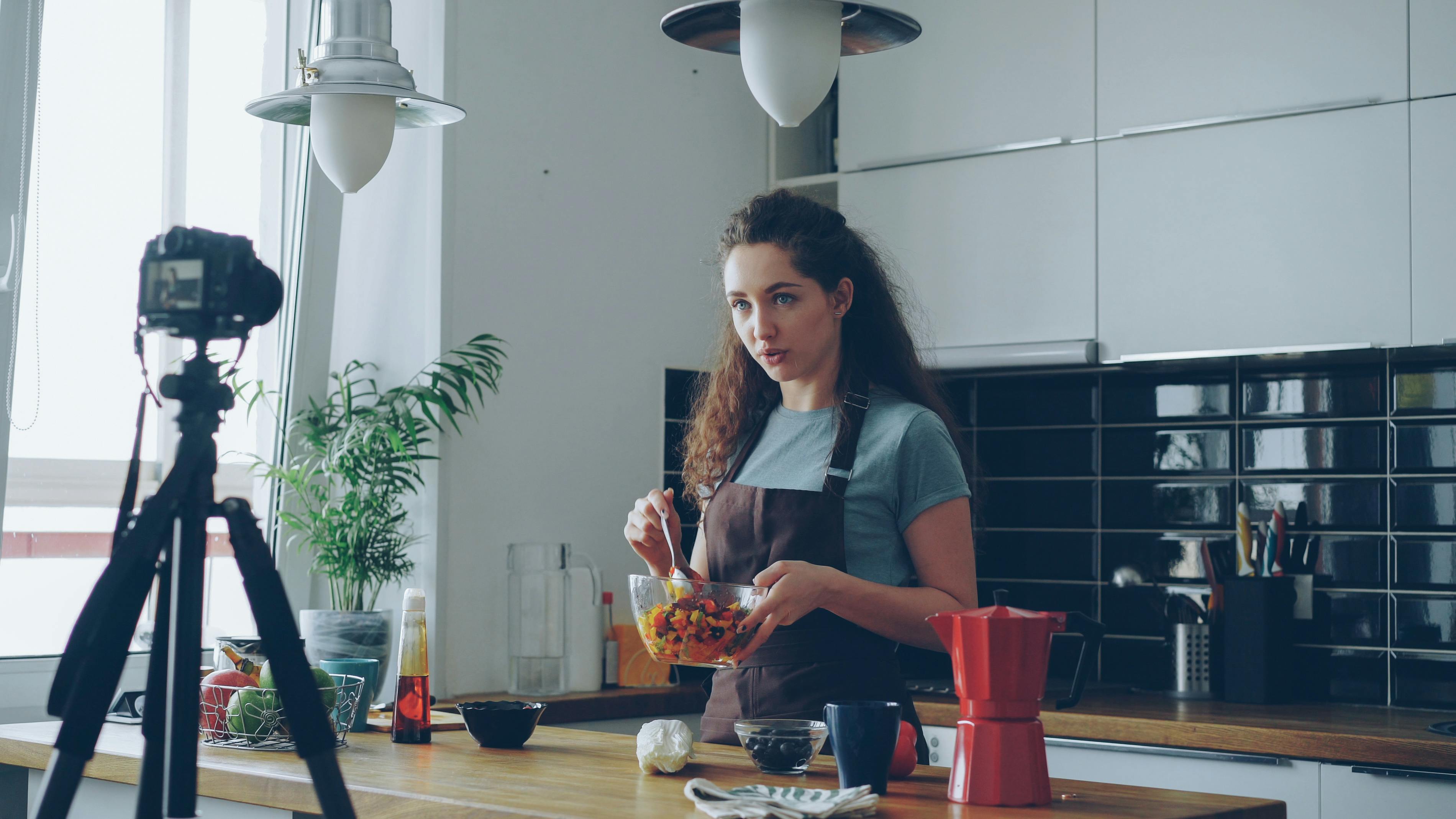 A woman records a cooking tutorial in her modern kitchen using a camera.