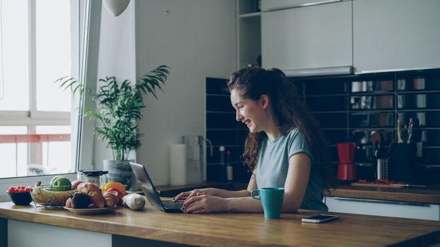 Young woman enjoys working on her laptop in a bright, modern kitchen setting.