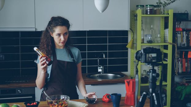 A woman filming a cooking vlog in her kitchen holding a condiment bottle.