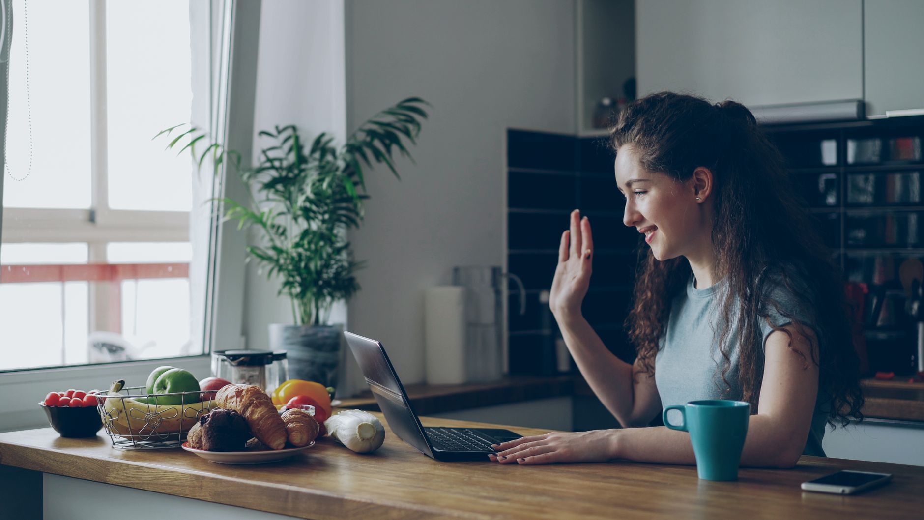 Woman waves during video call surrounded by healthy breakfast foods in modern kitchen.