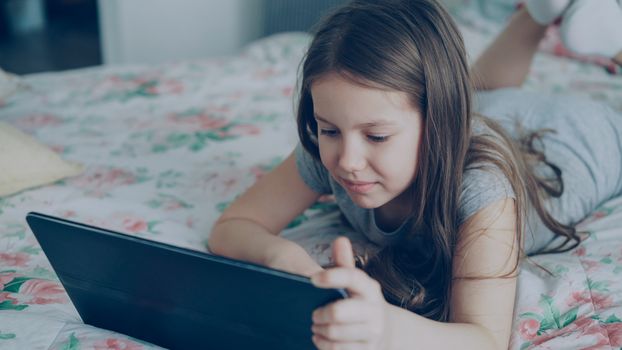 A young girl lies on a bed using a tablet, immersed in her digital world.