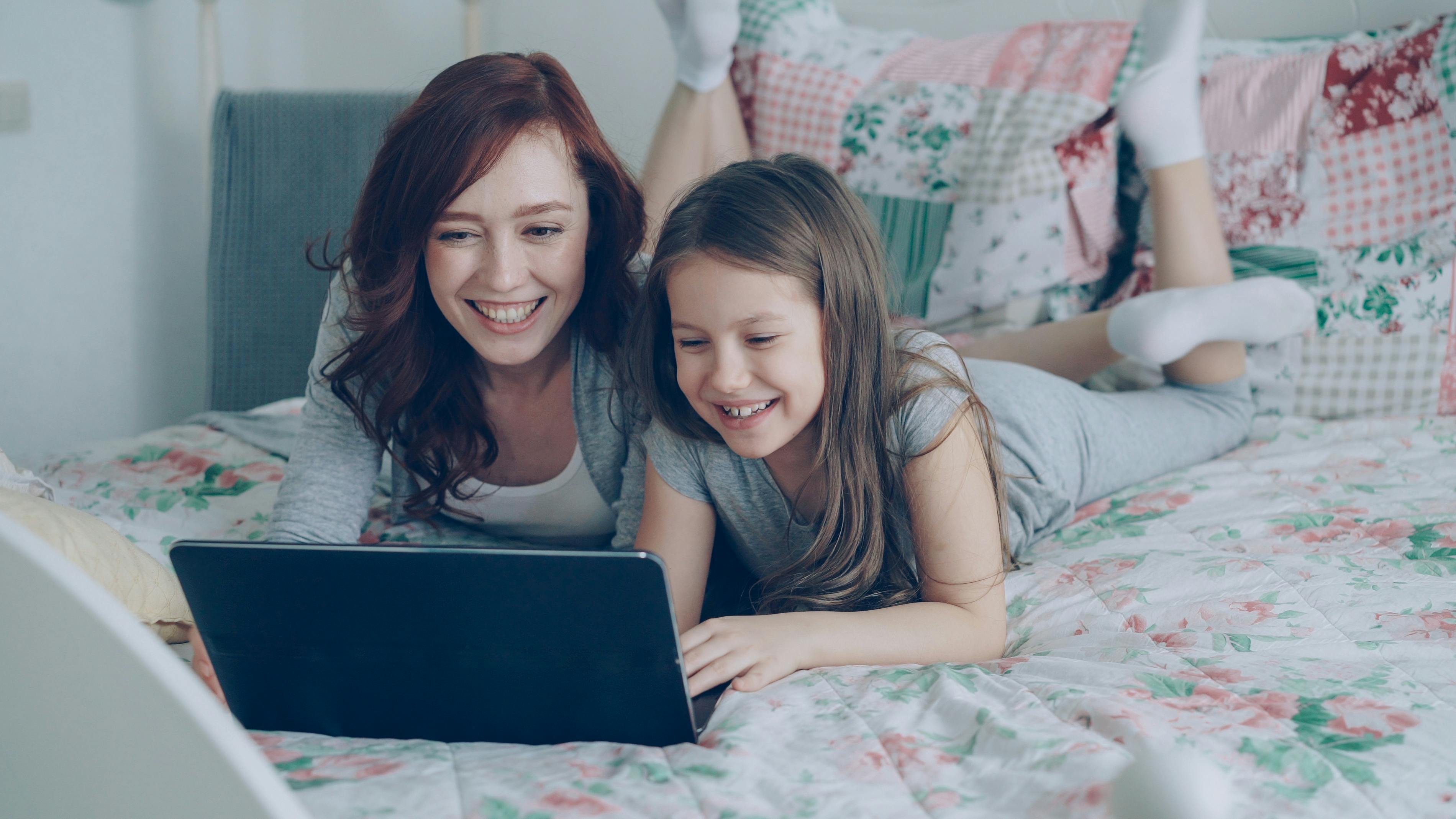 A joyful mother and daughter using a laptop together on a cozy bed at home.