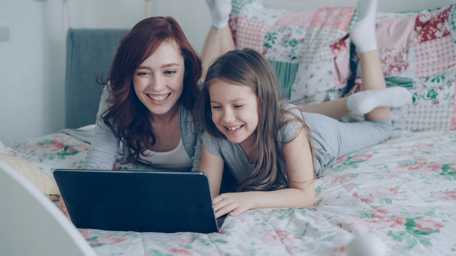 A joyful mother and daughter using a laptop together on a cozy bed at home.