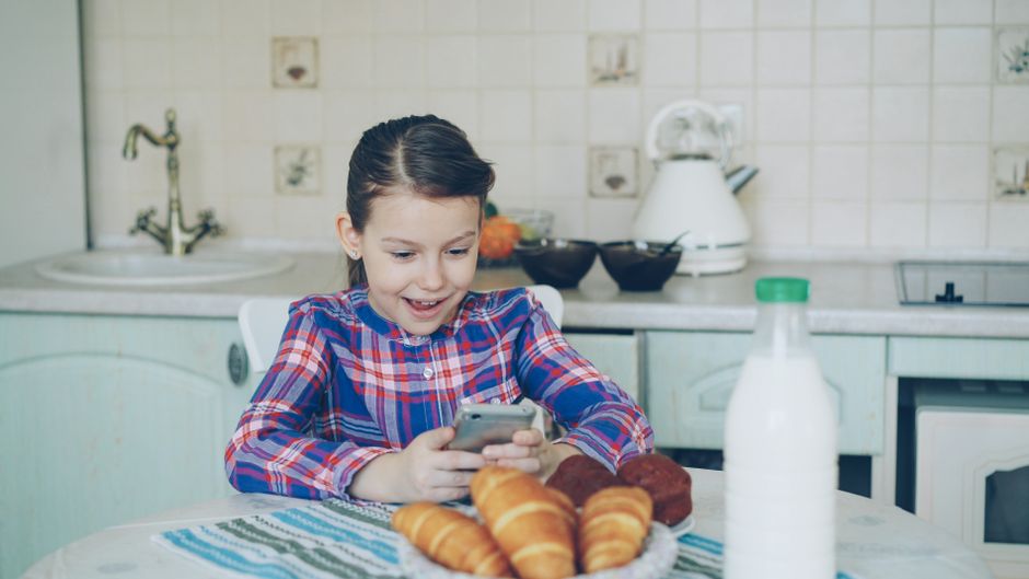Girl in kitchen using smartphone, smiling with breakfast pastries on table.