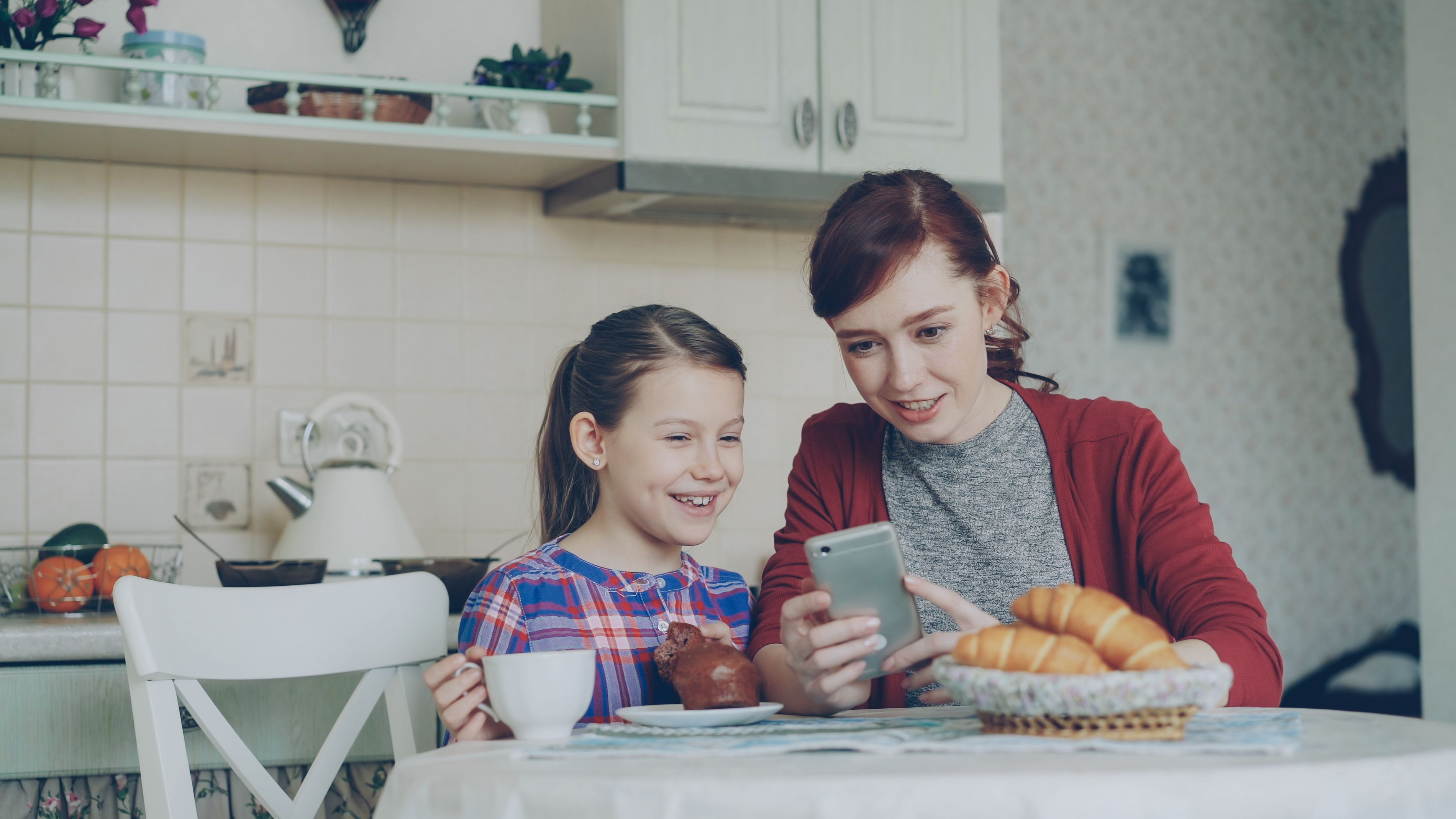 A mother and daughter smiling at a phone together in the kitchen