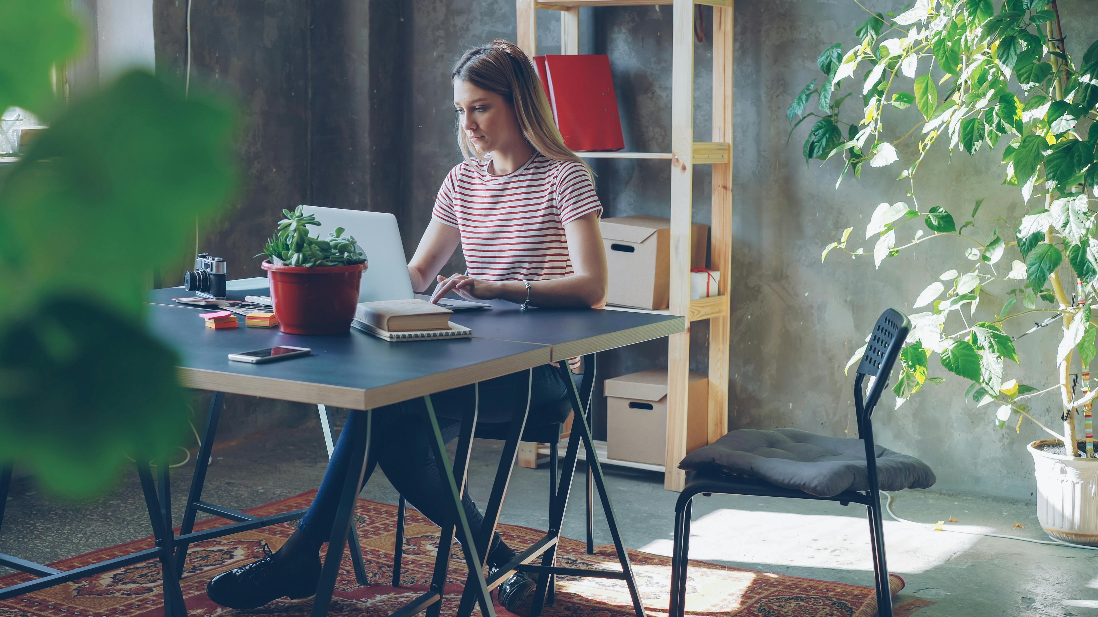 Woman in a striped shirt working on a laptop in a stylish home office with indoor plants.