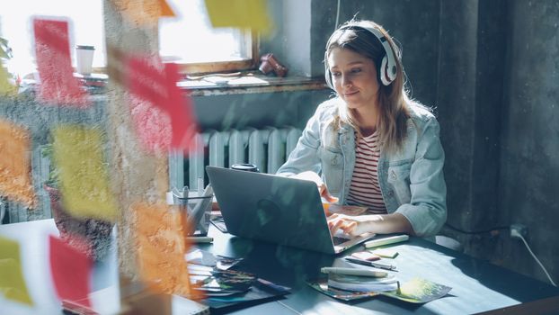 Young woman in casual attire working on a laptop with headphones at a creative workspace.