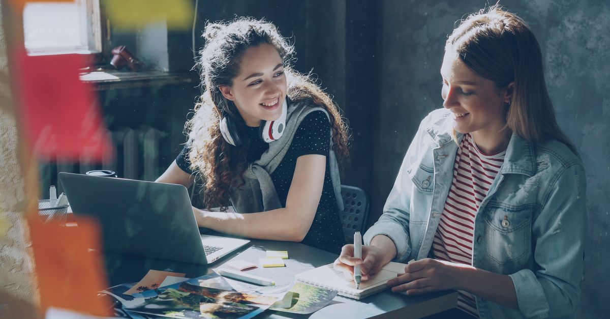 Two young women smiling and working together in a creative indoor setting with a laptop.