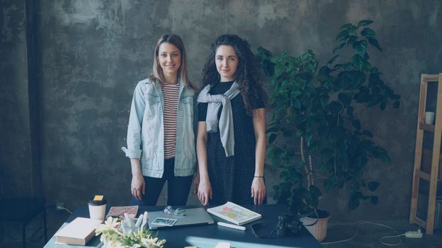 Two women in casual attire standing indoors by a desk, creating a cozy workspace vibe.