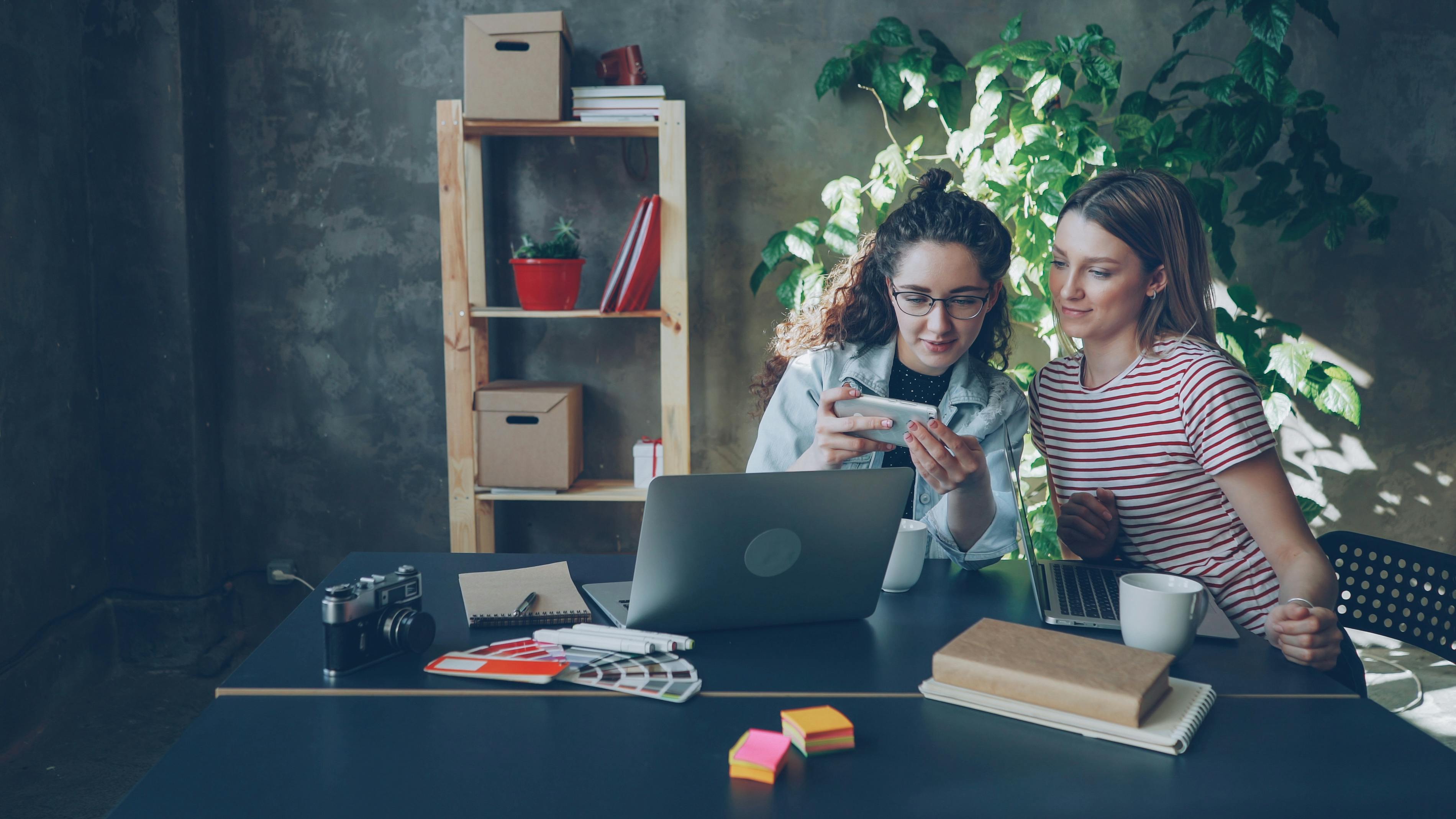 Two women working together in a cozy office setting, discussing ideas over laptops.