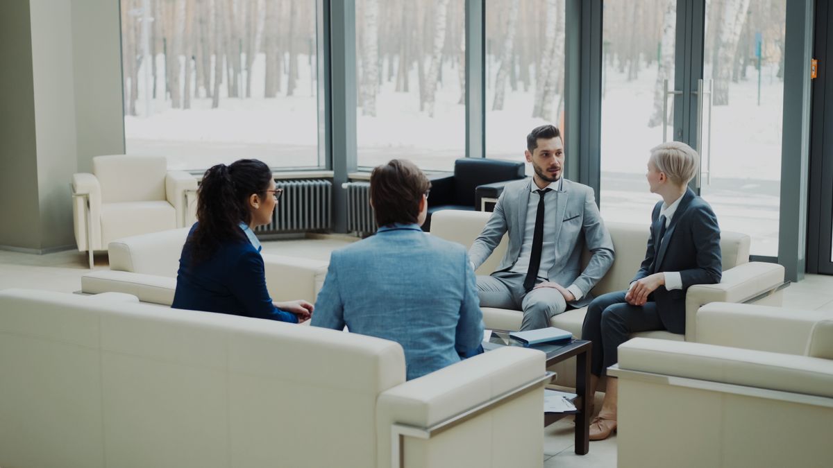 Four colleagues talking in a bright meeting lounge with birch trees outside the window