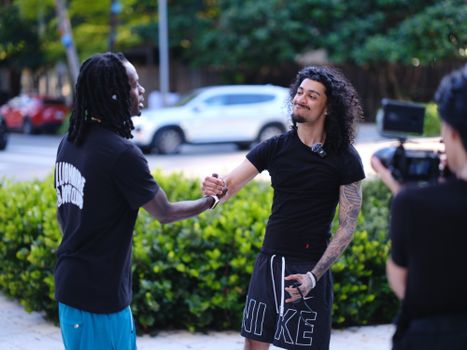 Two men greeting each other in an urban park setting while a camera records the moment.