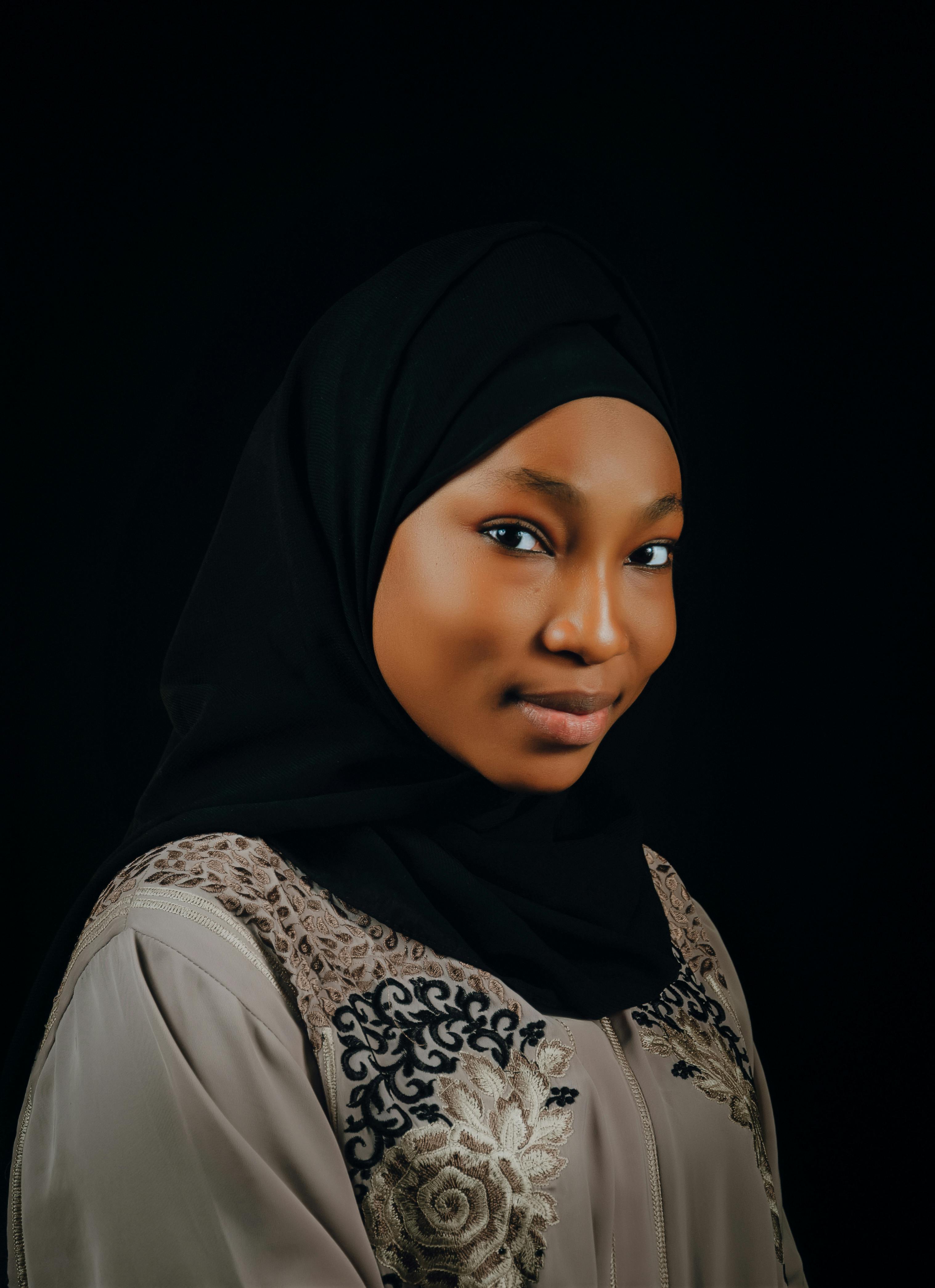 A woman in traditional attire and hijab against a dark background.
