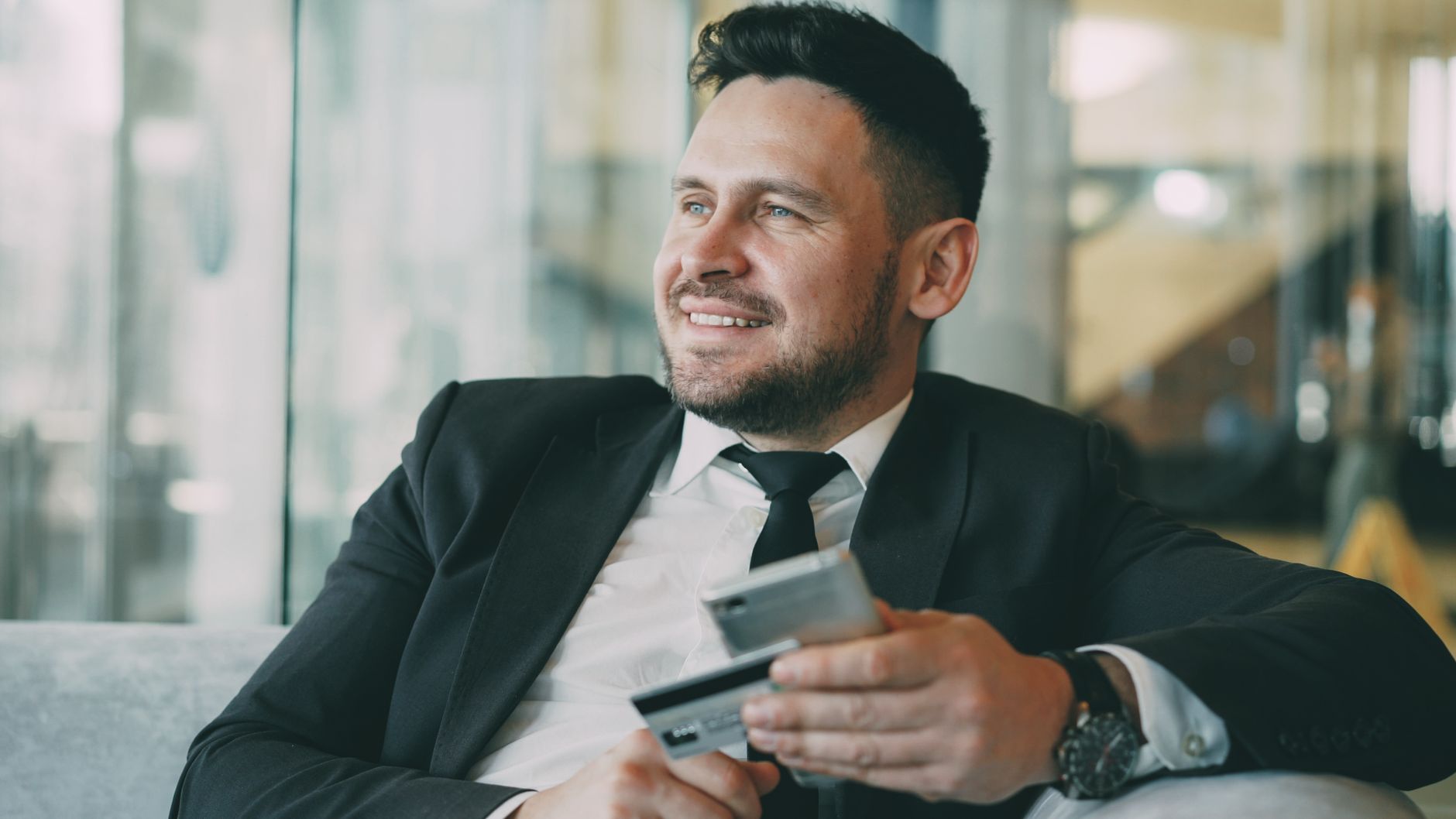 Smiling businessman in a suit holds a credit card and phone indoors, exuding confidence.