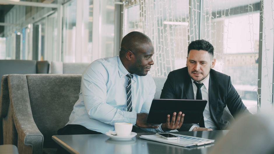 Two men in business attire collaborating over a tablet in a modern office setting. Two men in business attire collaborating over a tablet in a modern office setting.