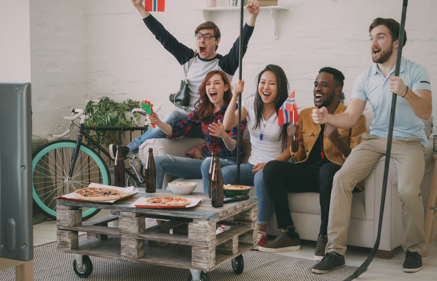 Excited friends cheering while watching a sports game on TV, holding flags and snacks.