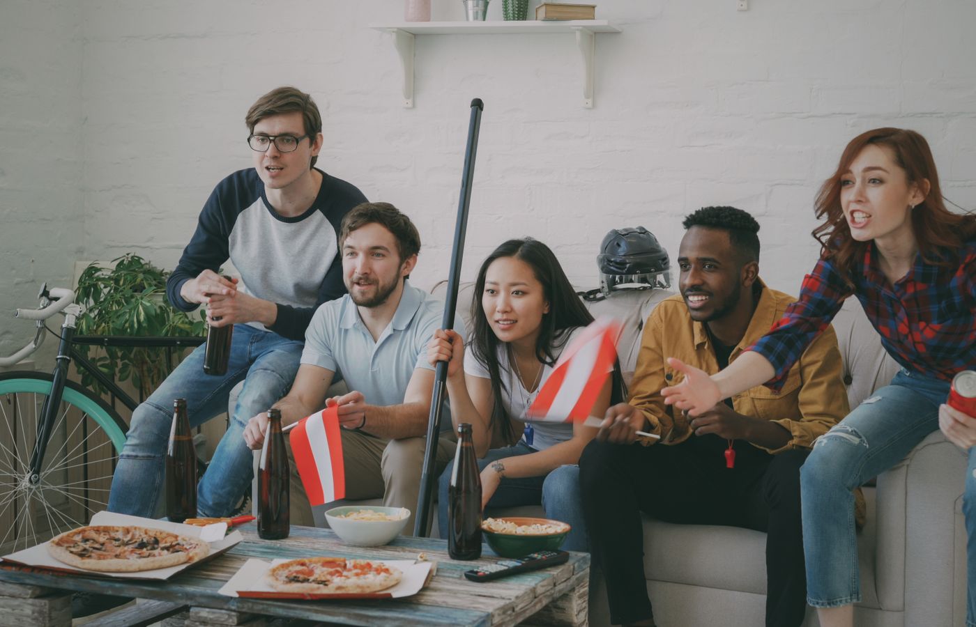 Group of diverse friends cheering for a game at home with snacks and flags.