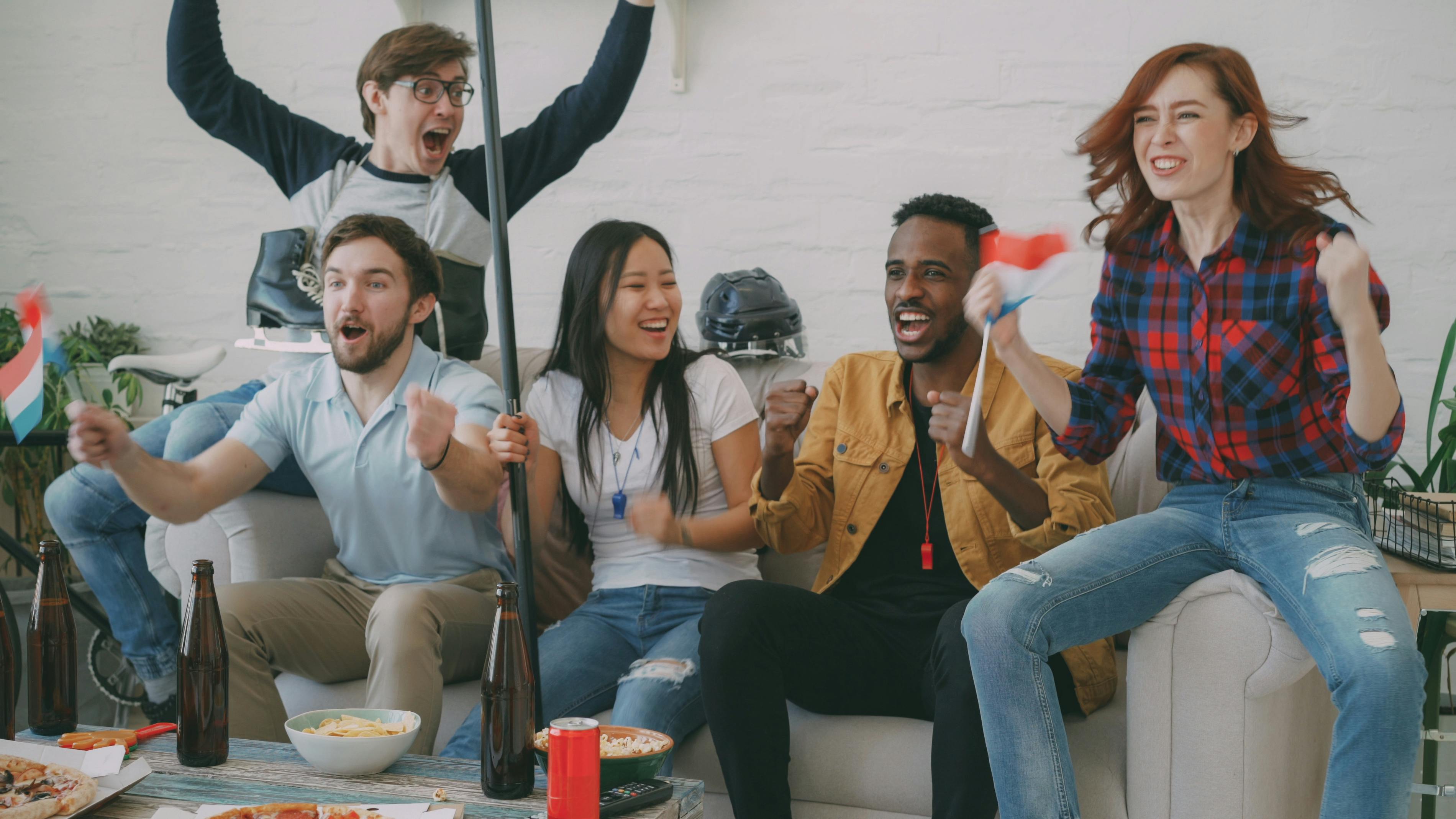 Group of diverse friends cheering indoors, holding flags, enjoying a sports victory celebration.