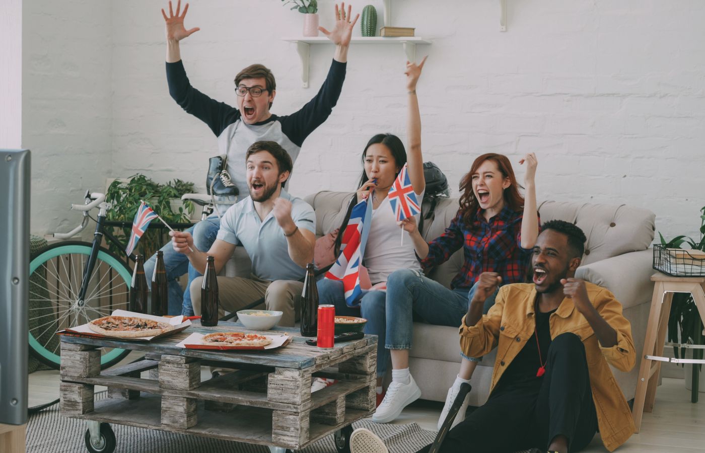 Friends joyfully watching sports on TV, waving flags, and celebrating indoors.