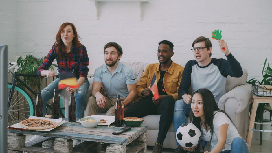 友人たちが室内でサッカーの試合を見ながら、旗を振ってスナックと飲み物で応援しています。