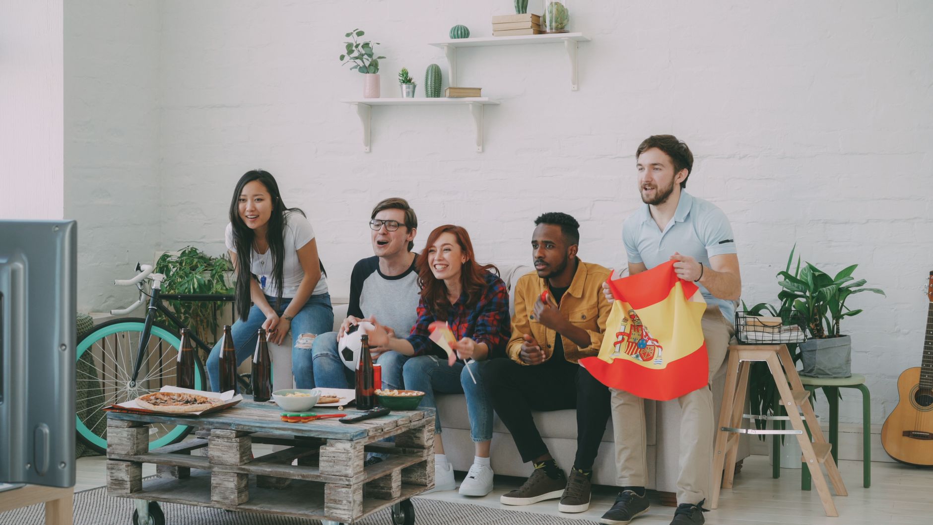 A group of friends eagerly watching sports on TV, holding a Spanish flag, indoors.