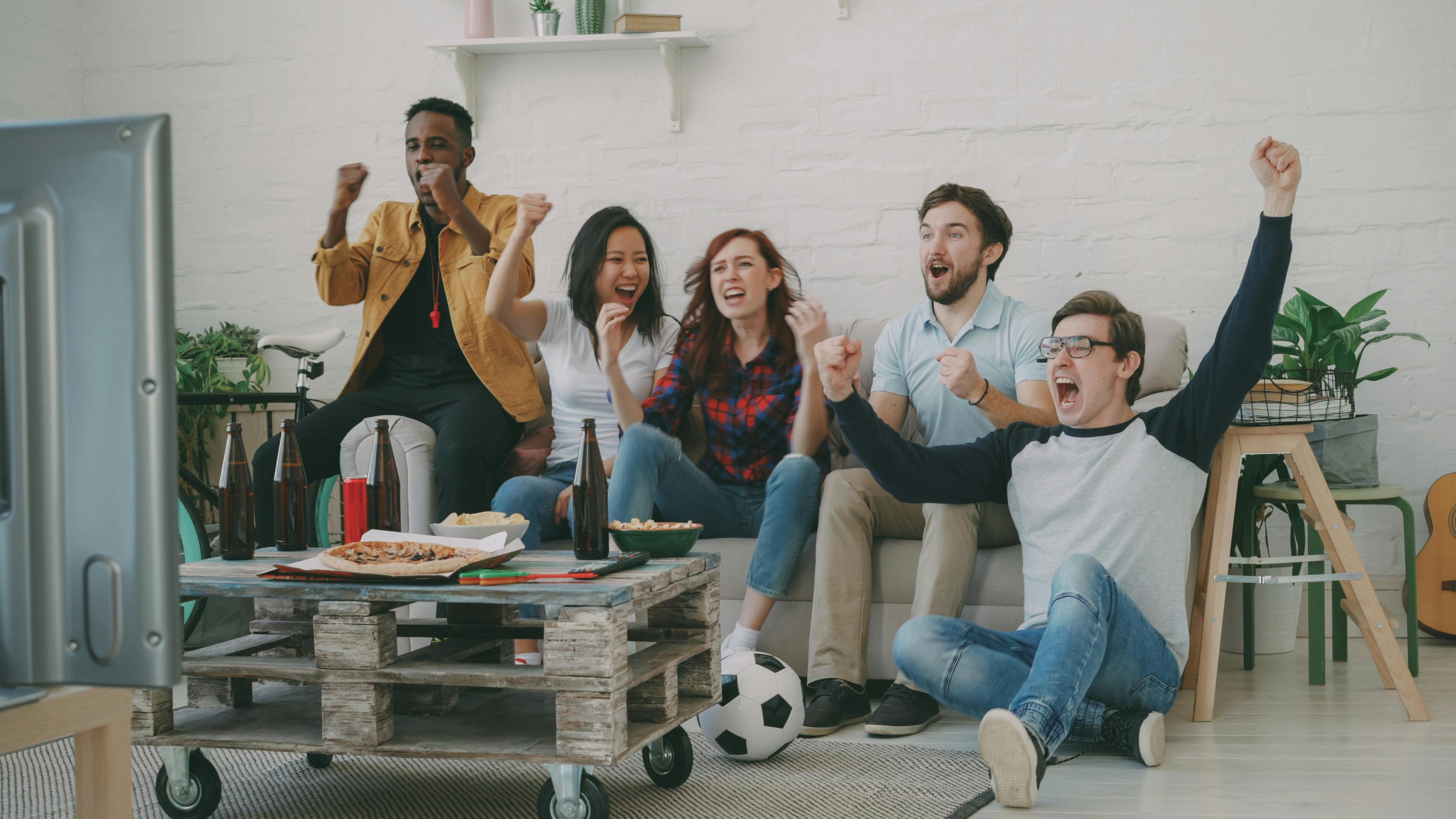 A diverse group of friends cheer excitedly while watching a game on TV at home.