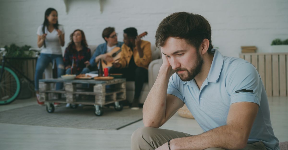 A young man sitting alone while others socialize in the background