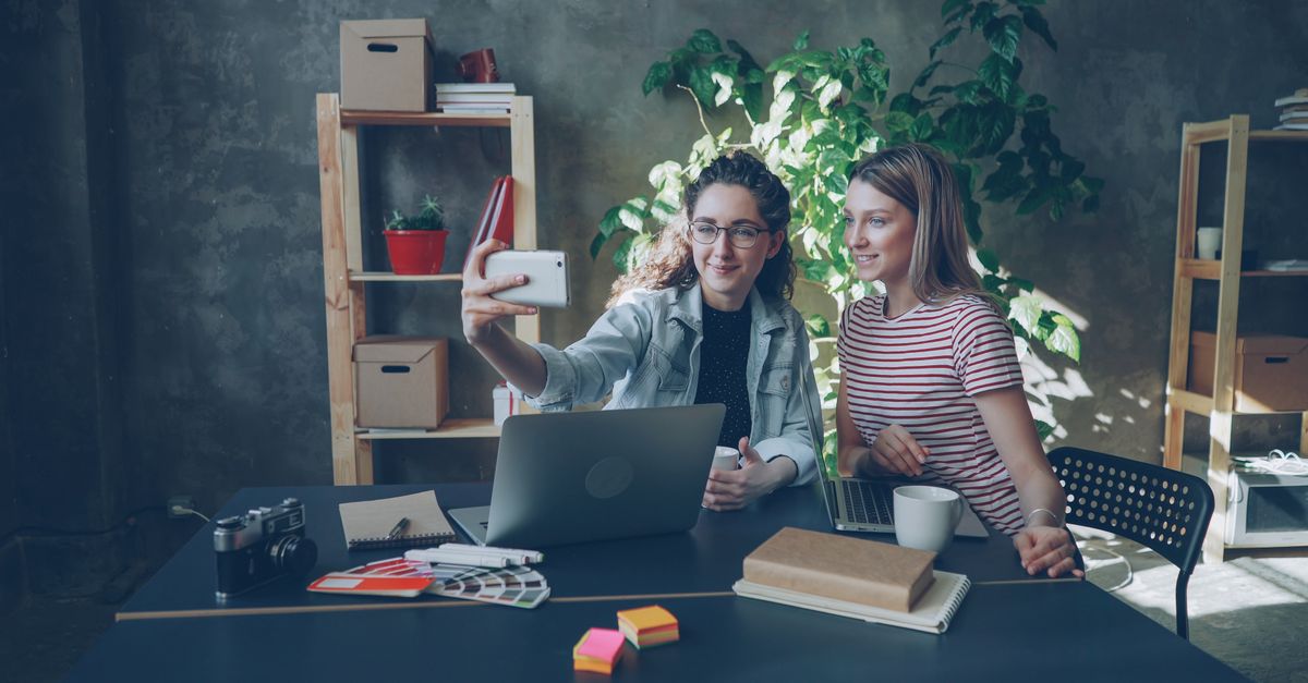 Two young women take a selfie together at their creative workspace, surrounded by laptops and plants.