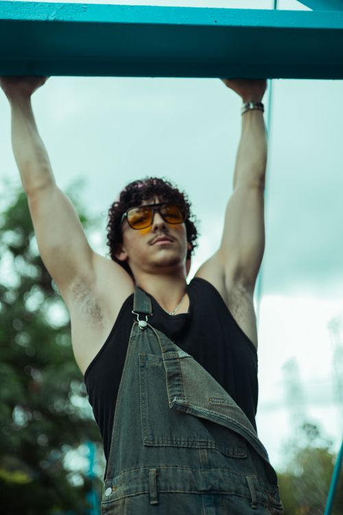 Free Adult man wearing overalls hangs from a beam outdoors, showcasing strength and casual style. Stock Photo