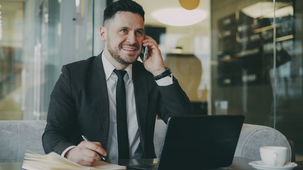 Professional man in a suit talking on the phone and working on a laptop indoors.