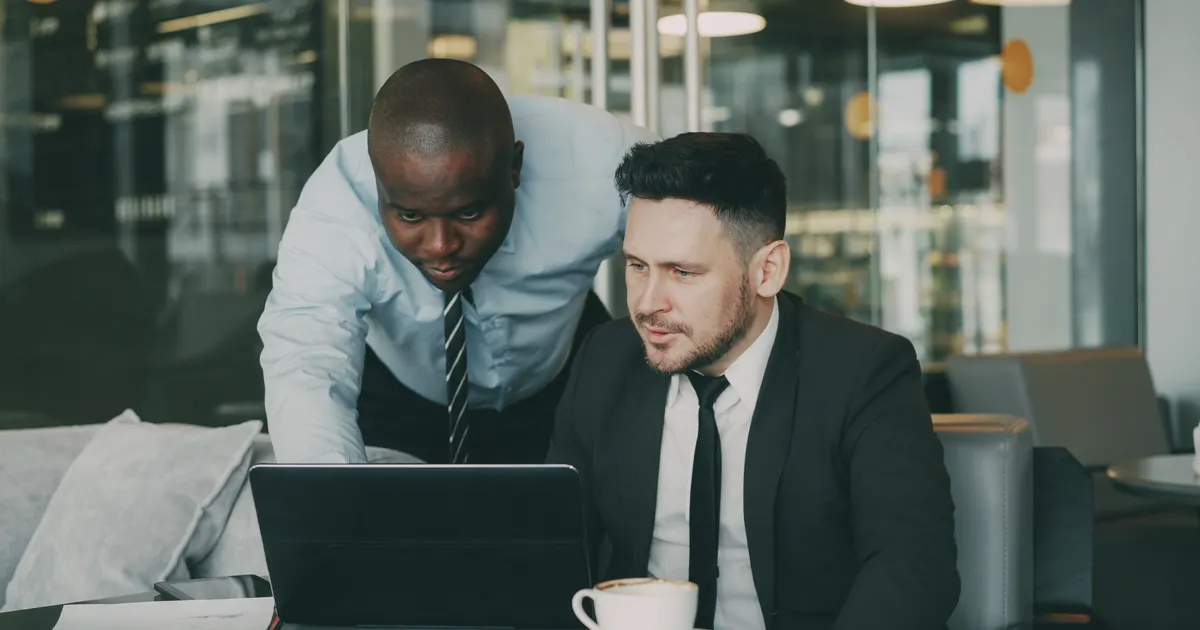 Two men in suits looking at a laptop on a table with a coffee cup, representing due diligence and compliance documentation workflows