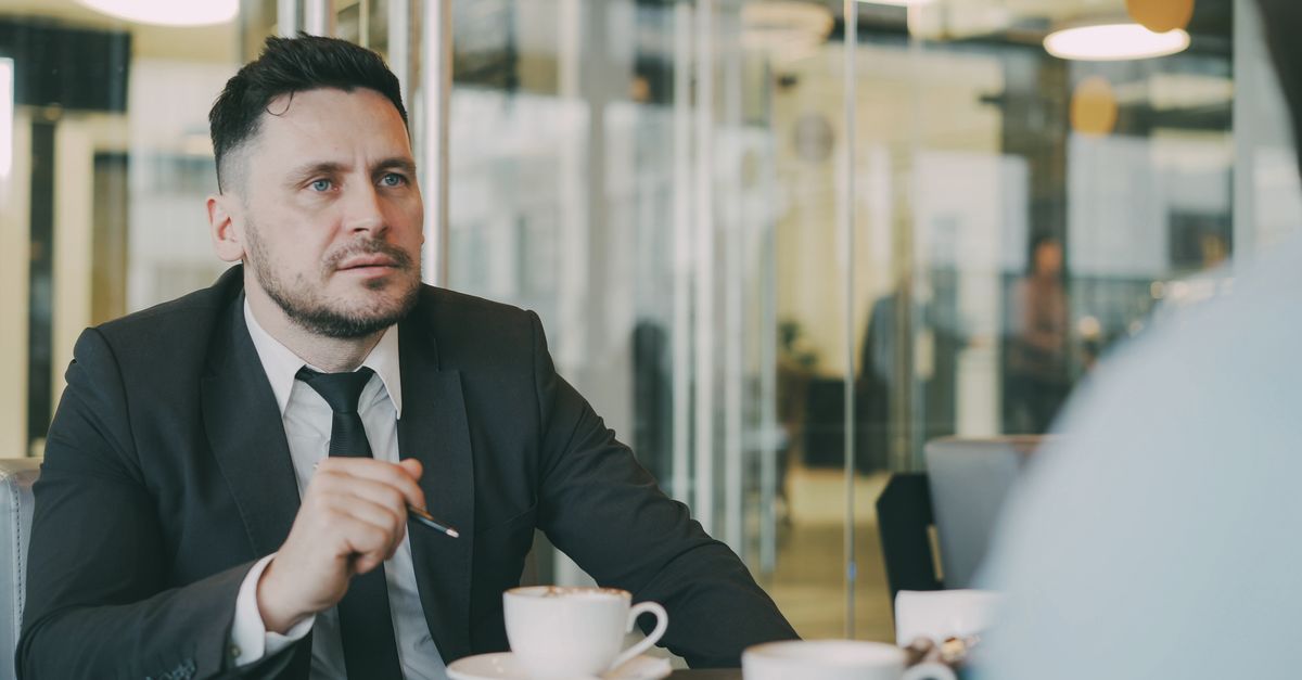 Man in suit holding coffee cup in modern office setting, engaged in conversation.