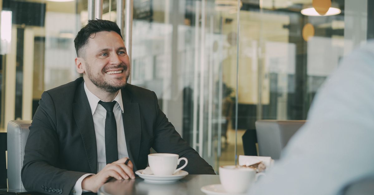 Professional businessman in a suit smiling during a coffee meeting in a modern office setting.