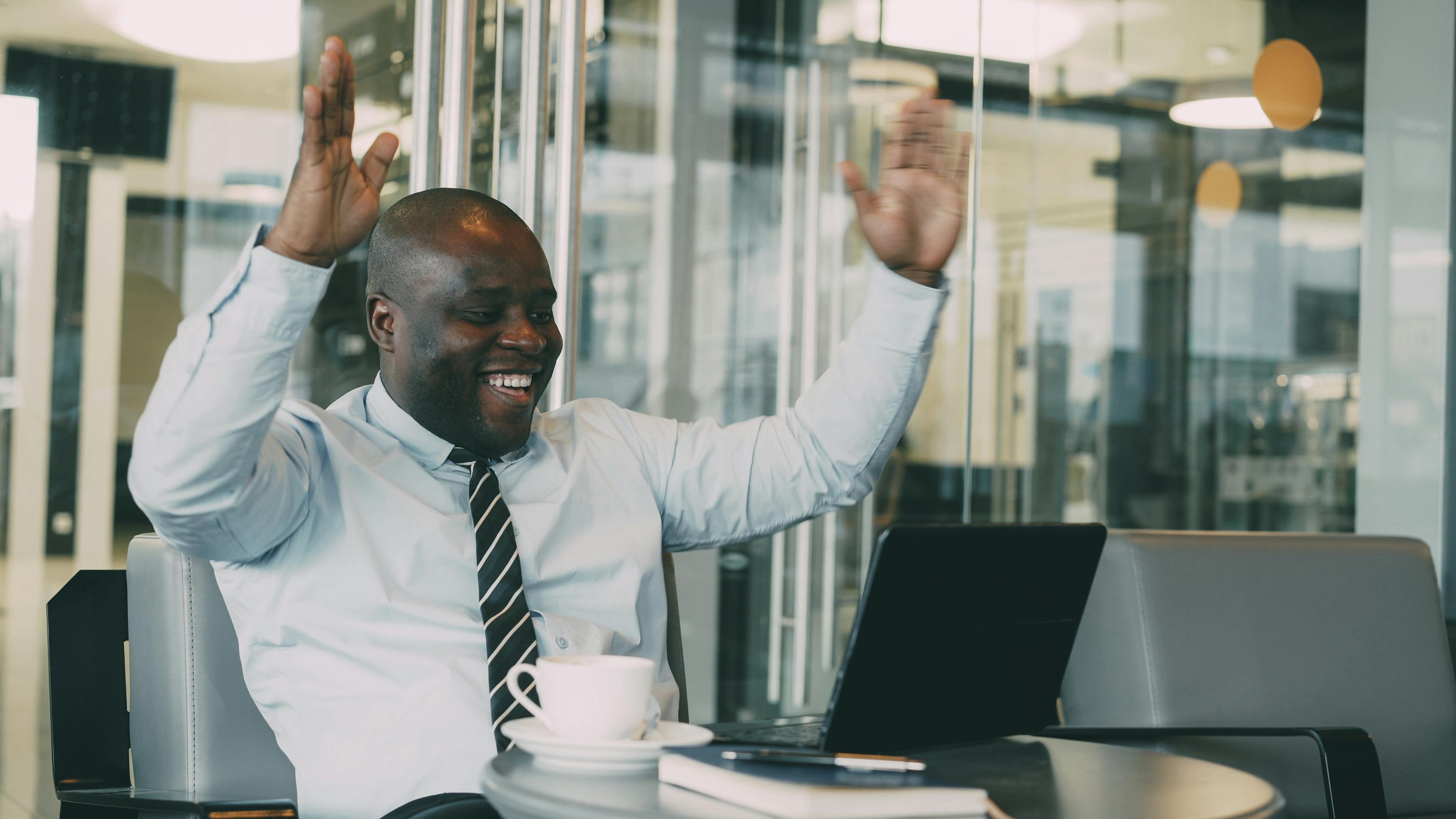 Smiling businessman raises arms in excitement at a cafe with laptop and coffee.