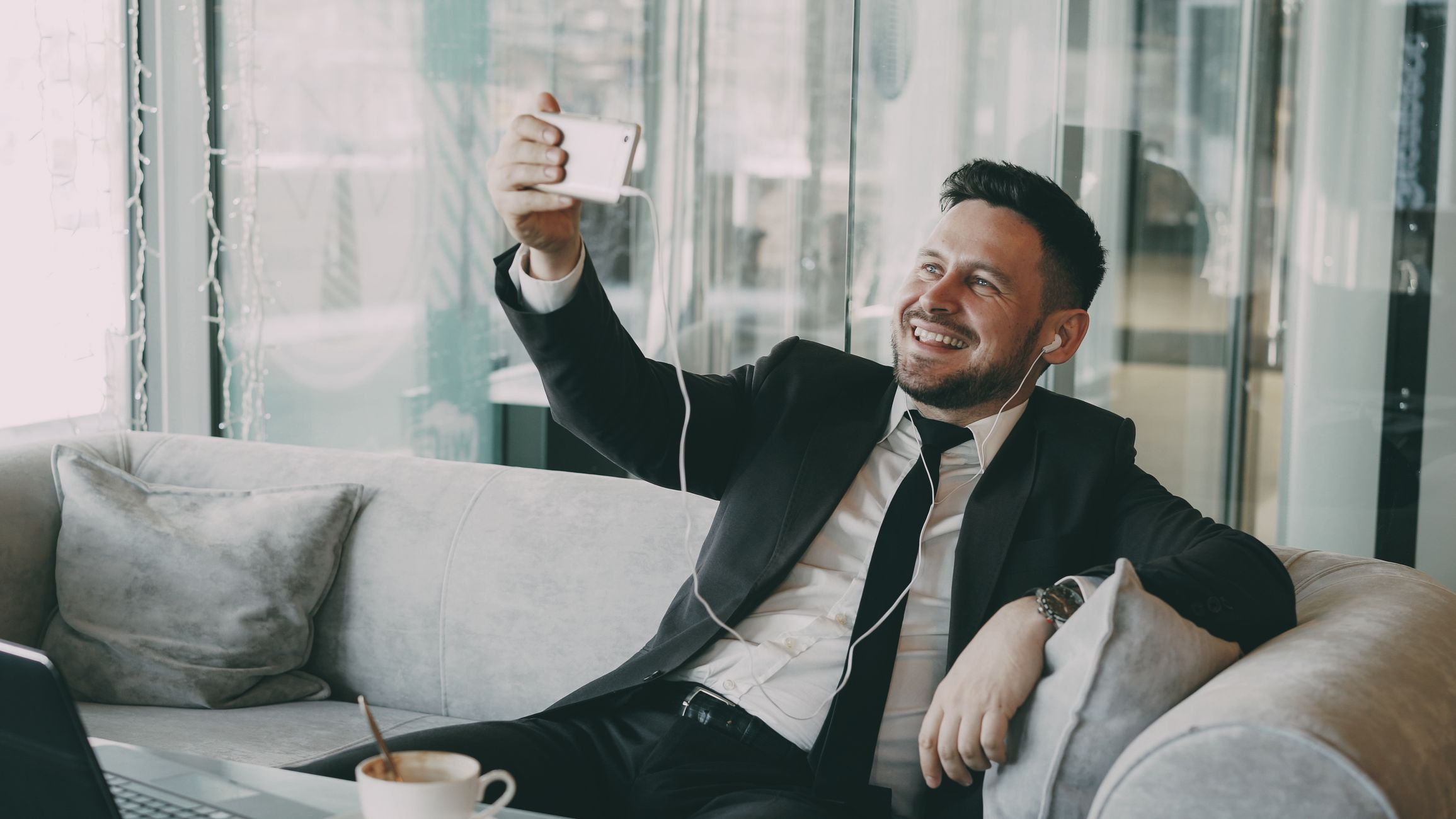 Smiling businessman in a suit taking a selfie while relaxing on a couch, capturing a candid moment.