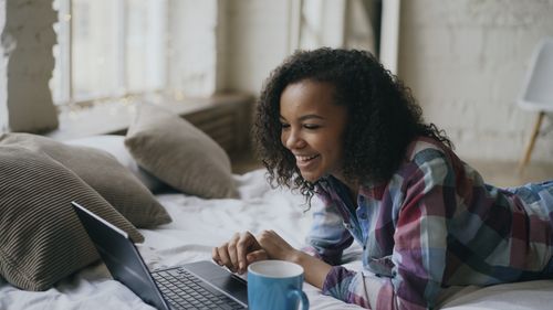 Free Happy woman using laptop on bed with a blue mug, enjoying leisure time indoors. Stock Photo