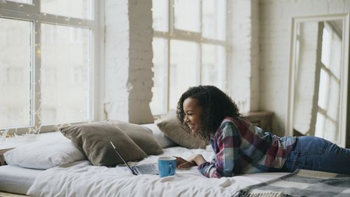 Free Woman lying on bed using laptop and drinking coffee in a cozy room. Stock Photo
