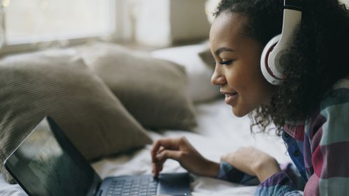 Free Smiling woman with headphones using laptop on bed. Comfortable home setting. Stock Photo