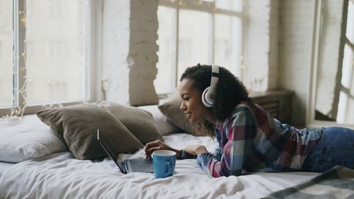 Free Woman lying on bed with laptop, headphones, and coffee, enjoying leisure time. Stock Photo