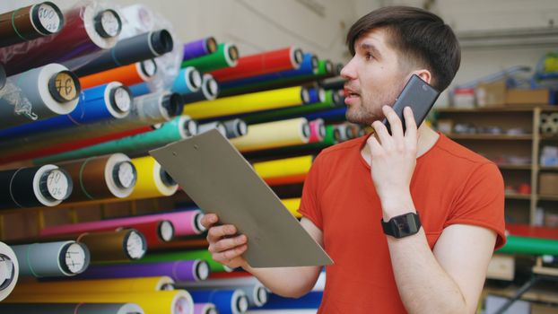Man talking on phone with clipboard in vibrant textile store, organized fabric rolls in background.