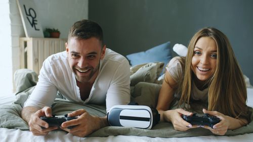 Free Young couple laughing and gaming in bed, with VR headset nearby, enjoying leisure time. Stock Photo