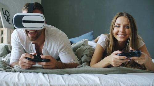 Free A couple playing video games together, one using VR glasses, in a relaxed bedroom setting. Stock Photo