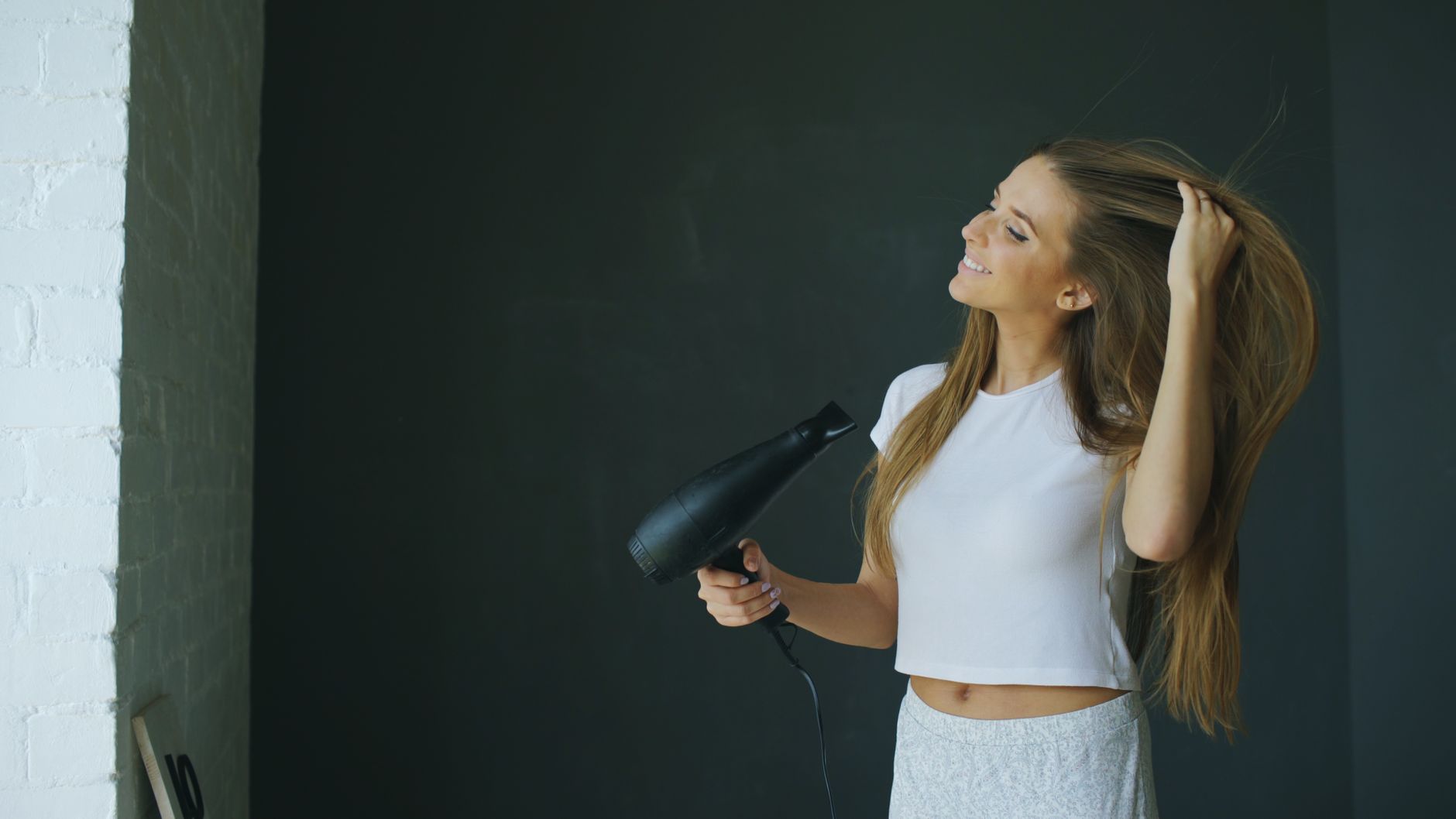 Young woman smiling while using a hairdryer for hair styling indoors.