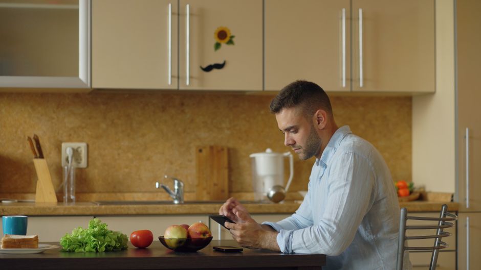 Man browsing smartphone in a modern kitchen with fresh produce on the table.