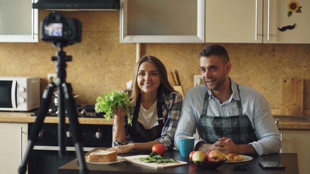 Couple recording a cooking video in their kitchen with fresh vegetables and fruits, smiling at the camera.